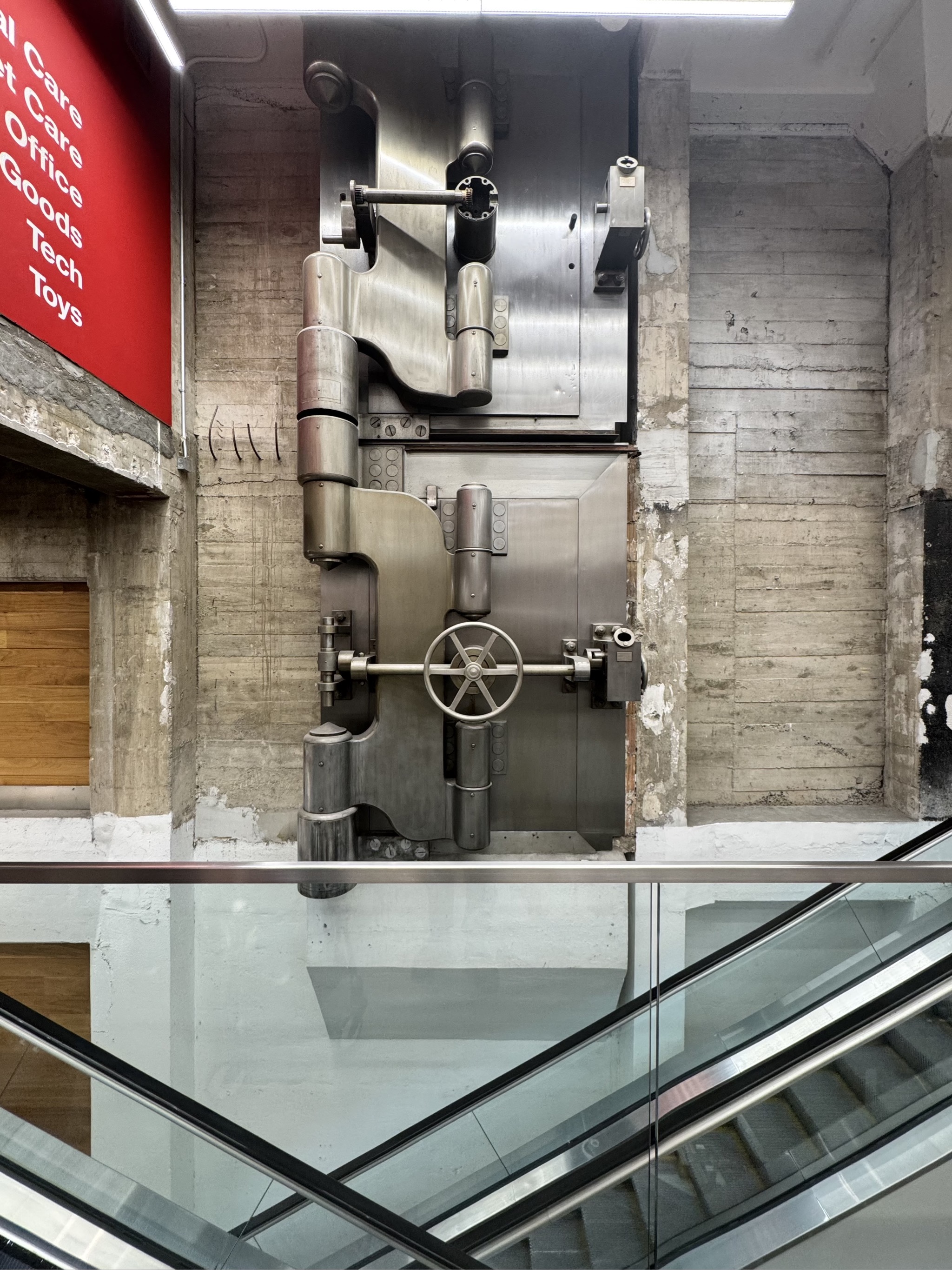 Massive bank vault door displayed in modern museum with glass railings.