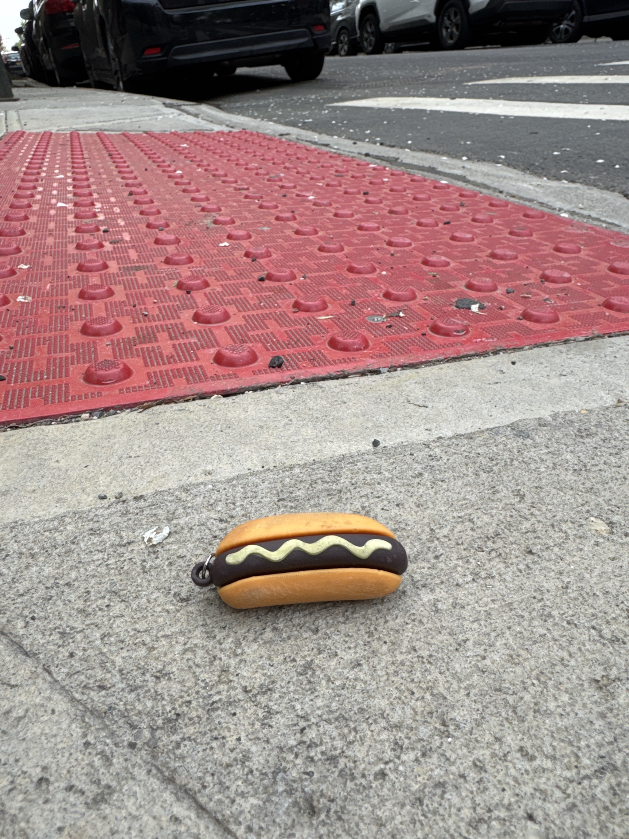 Hot dog keychain lies on sidewalk near red tactile mat.