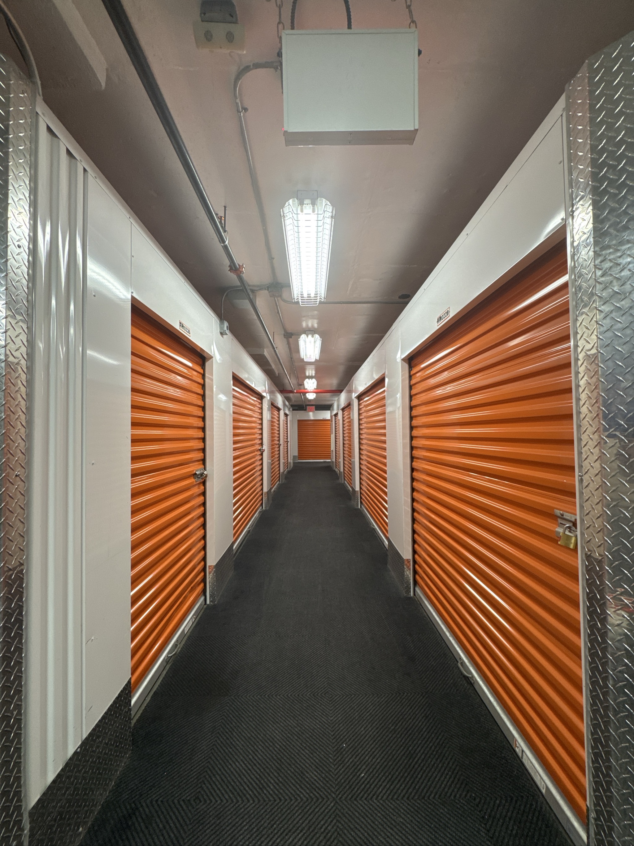 Indoor storage facility hallway with orange roll-up doors and fluorescent lighting.